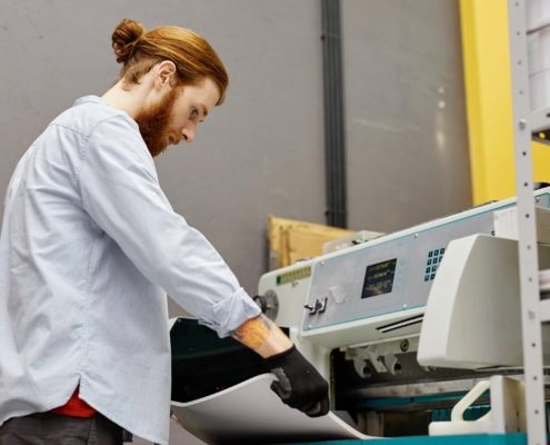 Side View Portrait Of Young Man Checking Quality Of Bulk Prints In Industrial Printshop
